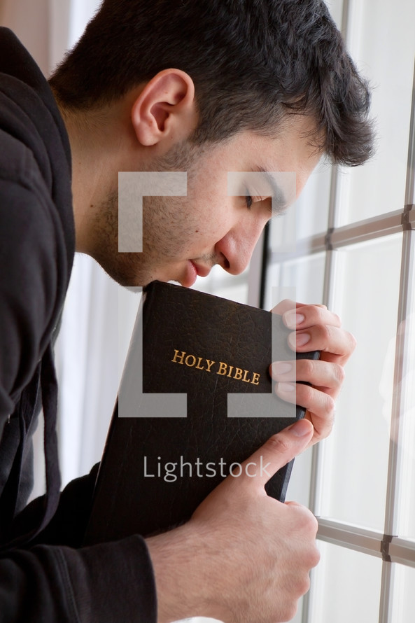 man praying by a window holding a Bible