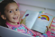 Girl in pajamas reading the Bible and smiling. She's reading the story of Noah before bedtime.  Reading a children's Bible.
