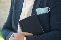 a man holding a Bible with a face mask in his pocket 