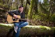 Man sitting on a fallen tree in the woods playing his guitar.