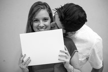 a woman holding up a blank sign and a man kissing her on the cheek 
