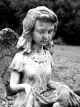 A statue of a female angel figure in a garden or graveyard setting. A black and white image of a female Angel weathered by storms, age, and life as a reminder that we have Gods angels looking over us as a constant reminder that this is not our permanent home. 