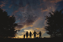 silhouette of a family standing outdoors holding hands under an evening sky 