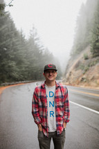 a man standing on the side of the road wearing a ball cap and plaid shirt 