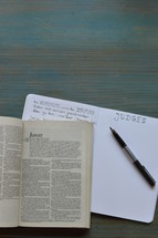 bible studies with a bible open at the book of Judges with notes and pen on a wooden cyan desk