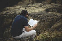 a man sitting on a rock outdoors reading a Bible 