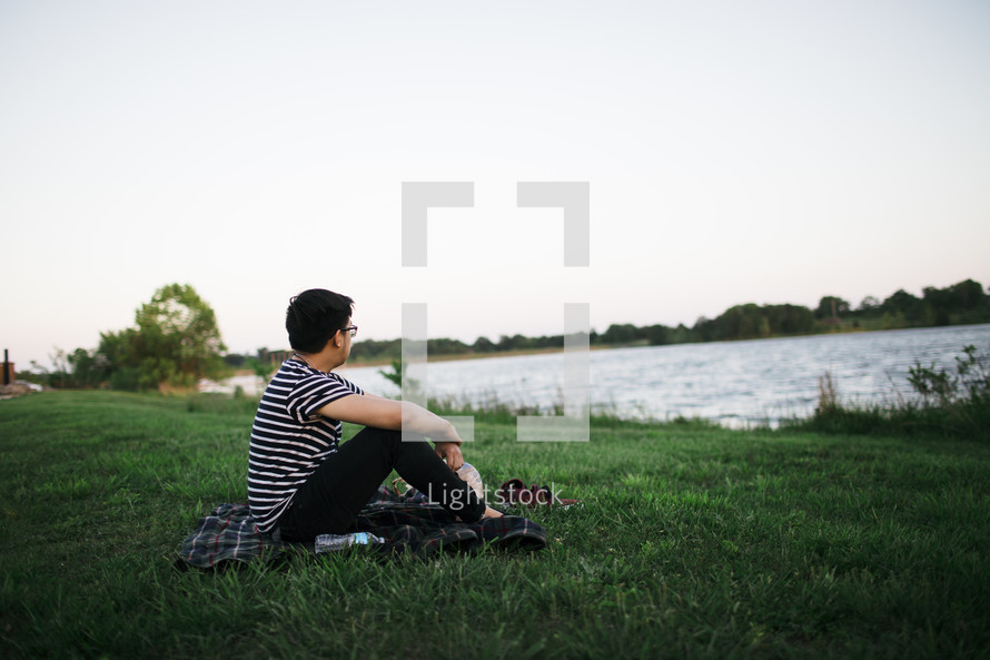 man sitting on a blanket in the grass by a lake 