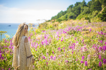 A blonde woman in a field of flowers next to the ocean.