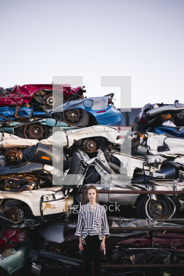 a woman in a scrap metal junk yard standing by crushed cars 