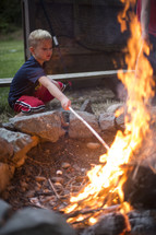 A young boy poking a campfire with a stick.