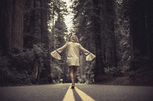 A woman walks along the center of a highway through a forest.