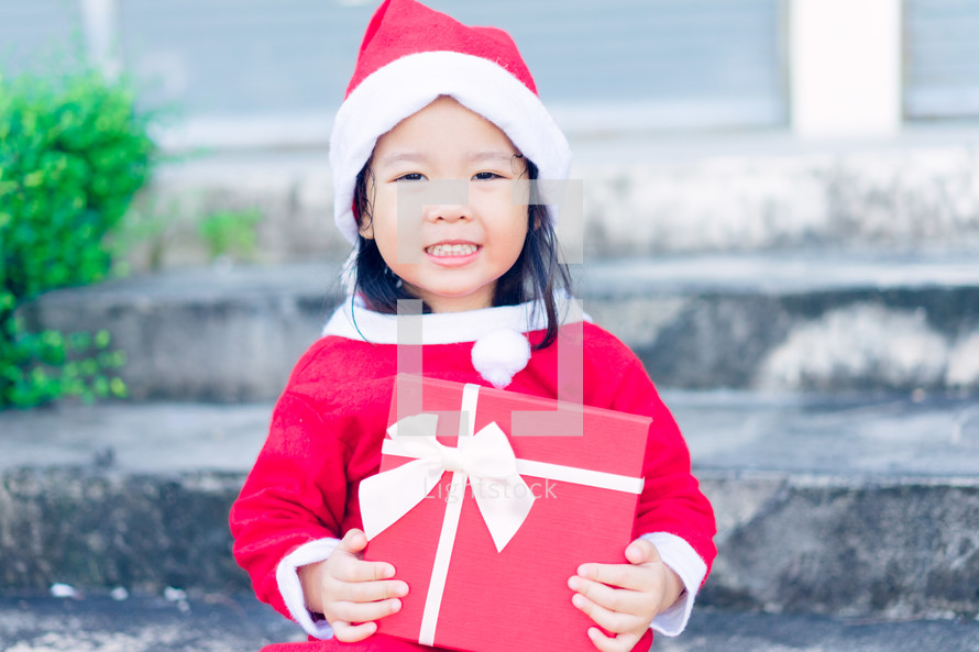 a little girl in a Santa suit holding a red gift box 