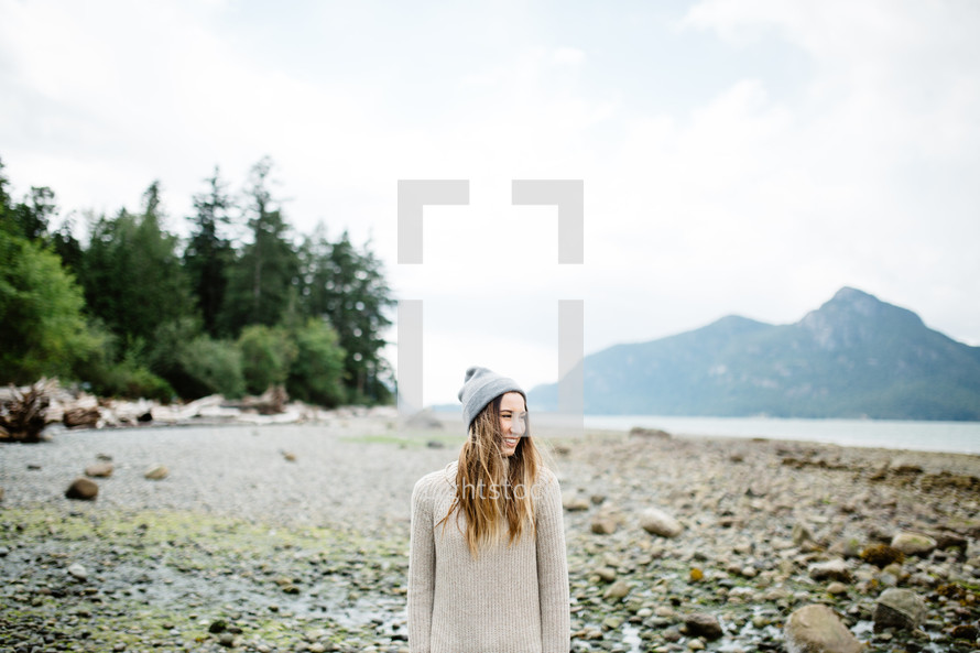 woman in a wool cap standing on stones on a shore 