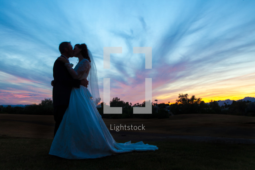 Bride and groom kissing, standing on a hillside with a sunset behind them