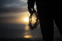 a man holding a crown of thorns on a shore at sunset 