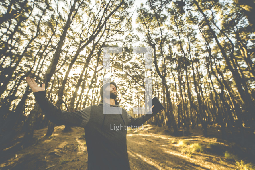 a man with outstretched arms holding a Bible looking up to God in a forest 