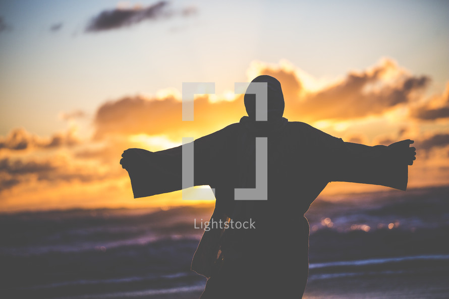 silhouette of Jesus with open arms on a shore at sunset 
