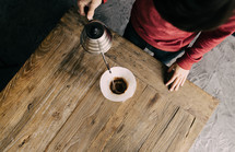 A man making coffee by pouring it over a filter in a chemex on a wooden plank table.