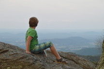 a boy sitting on a mountaintop taking in the view 