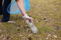 A woman hands in gloves collects and puts used plastic bottle into a blue trash bag. A volunteer cleans up the park on a sunny bright day. Clearing, pollution, ecology and plastic concept.