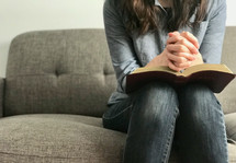 A woman sitting on a sofa praying with her Bible
