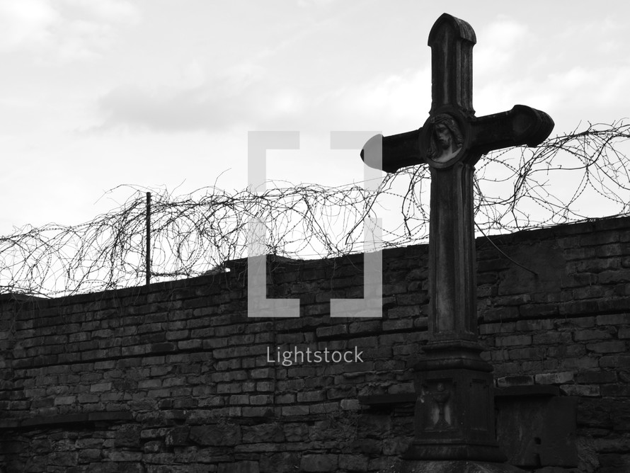 Stone cross in front of a wall with a barb wire fence.
