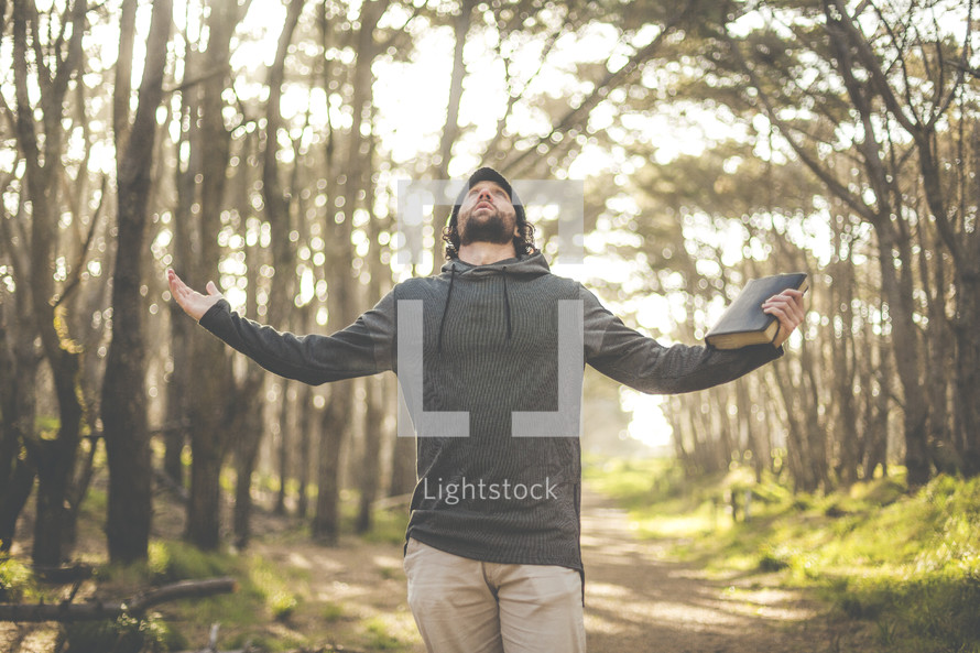 a man holding a Bible with outstretched arms looking up to God in a forest 