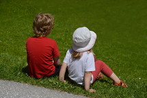 two little children sitting in the grass and watching something far away. 
