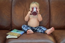 toddler boy sitting on a couch sipping from a mug 