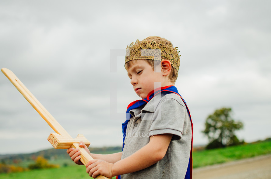 A young boy wearing a cape and crown  holds a wooden sword.