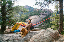 a woman camping sitting in a hammock next to her golden retriever 