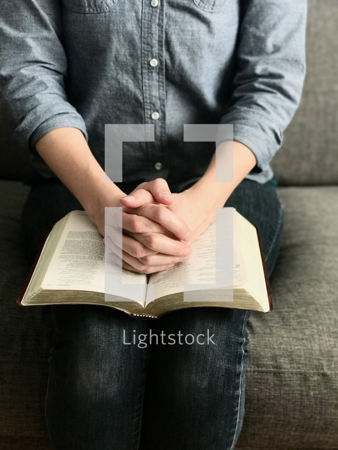 A woman sitting on a sofa with her hands clasped in prayer over an open Bible