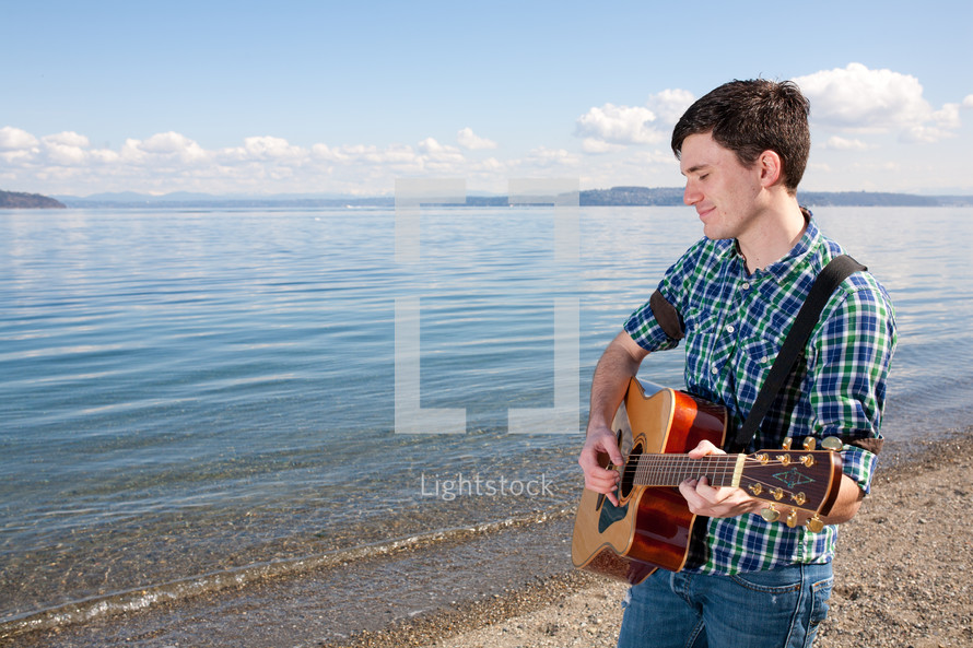 Smiling man playing guitar at water's edge.