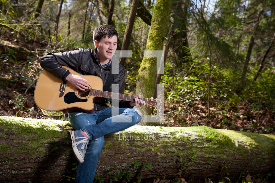 Man sitting on a fallen tree in the woods playing his guitar.