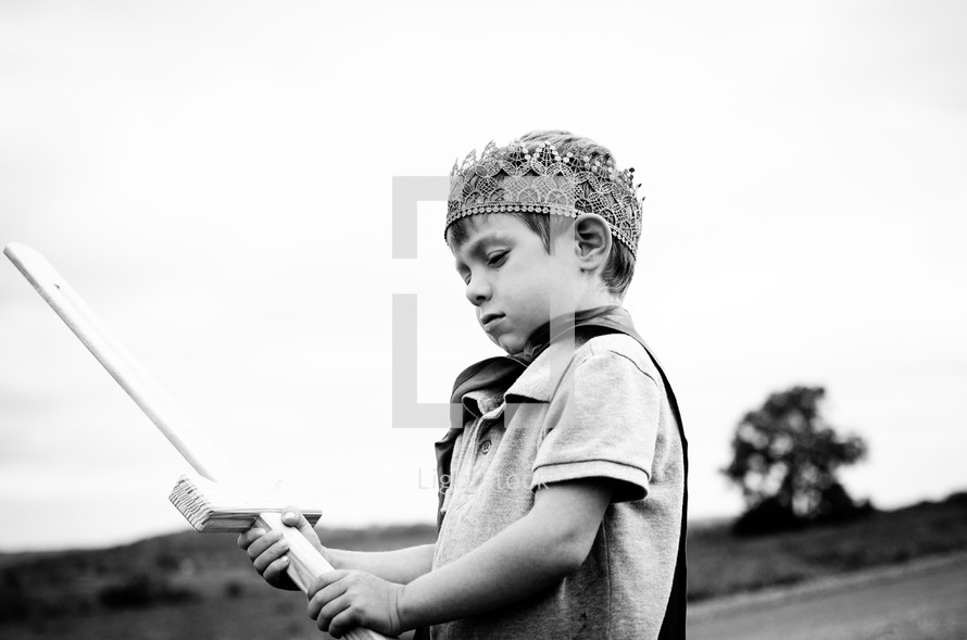 A young boy holds a sword outside wearing a cape and crown.