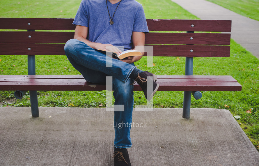 A man reads a Bible on a bench in a park.