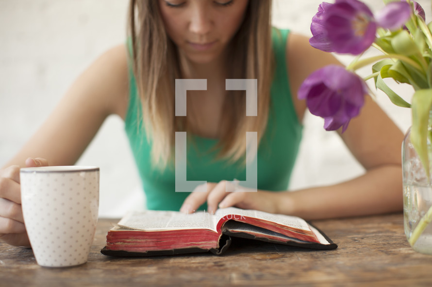 Woman with a coffee cup reading the Bible on a wood table with tulips.
