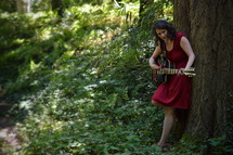 woman standing under a tree in a forest with a guitar 
