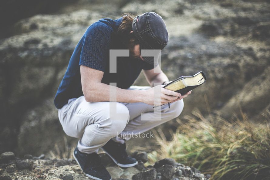 a man squatting with head bowed in prayer holding a Bible 