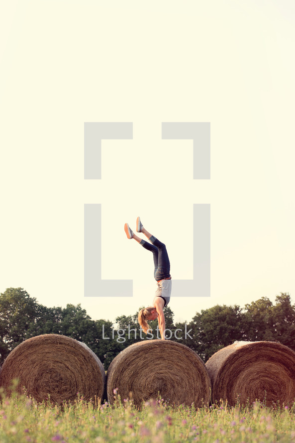 Woman doing a handstand on a rolled bale of hay in a field.