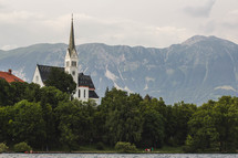 A church with a tall steeple surrounded by mountains and a lake.
