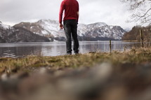 A man in a red shirt standing by a lake near a mountain range.