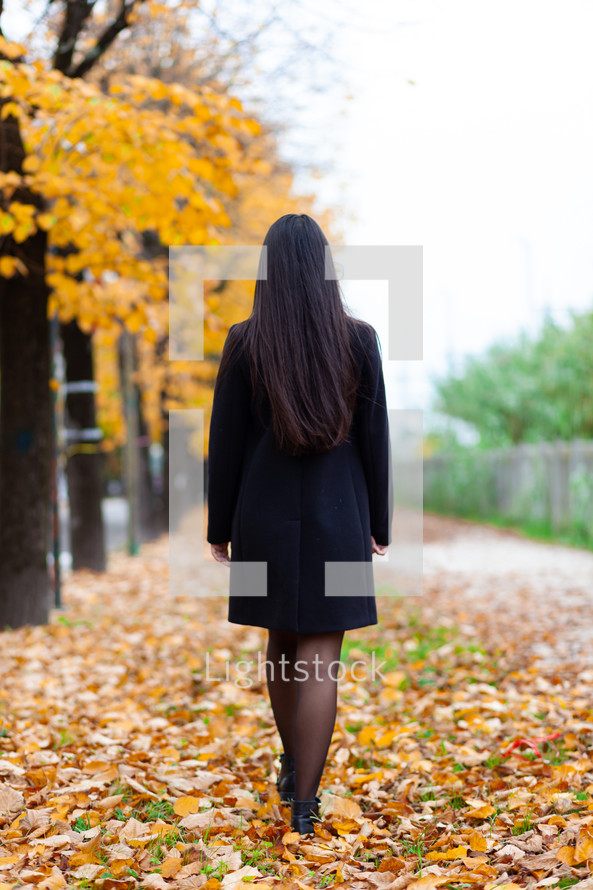 Young woman is walking away on a path in a park covered with yellow fallen leaves in autumn