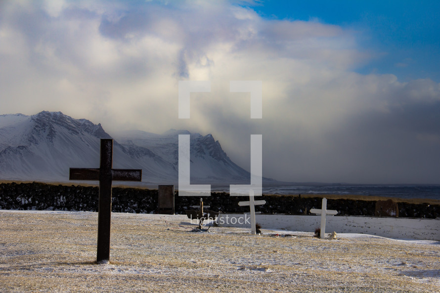 Cemetery in Iceland with several cross grave markers and dramatic mountains, clouds and sea in background