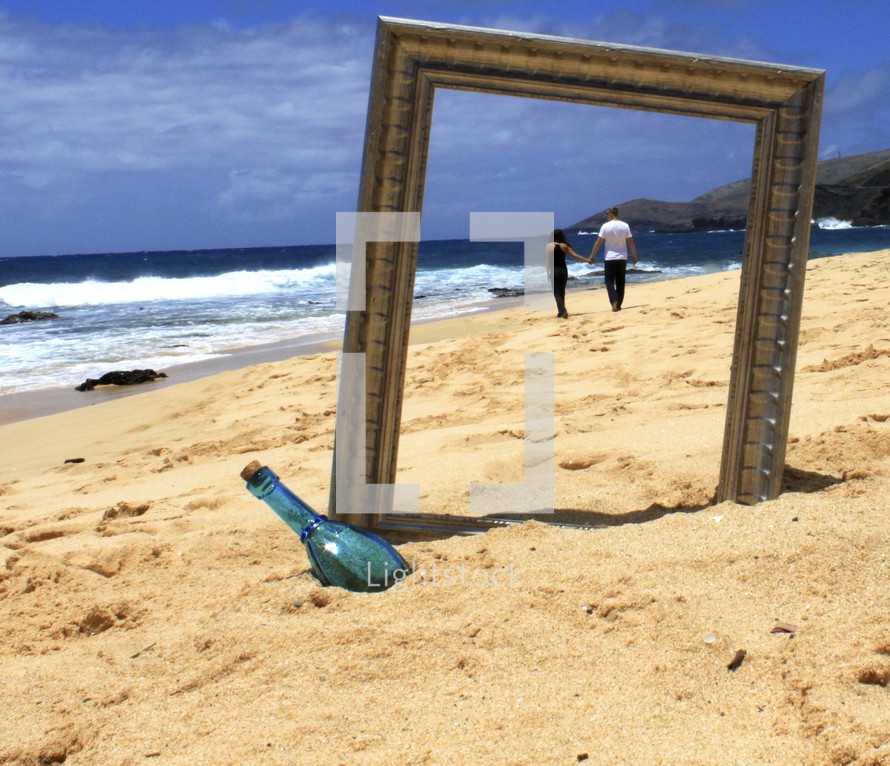 Couple holding hands while walking on the beach, seen through a frame near a glass bottle.