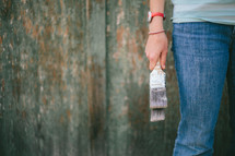 Leg and arm of a woman holding paintbrushes standing against a wooden wall