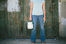 Woman standing in front of old wooden wall holding paint can and paint brush