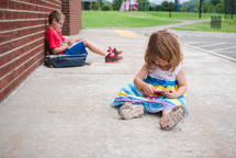 a boy doing his homework leaning against a school building and toddler sister playing 