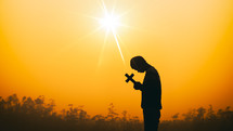 a male holding a cross at sunset against an orange sky