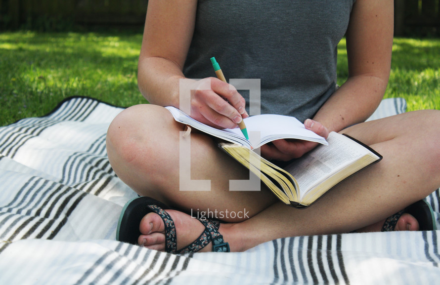 a girl sitting on a blanket in the grass reading a Bible and writing in a journal 